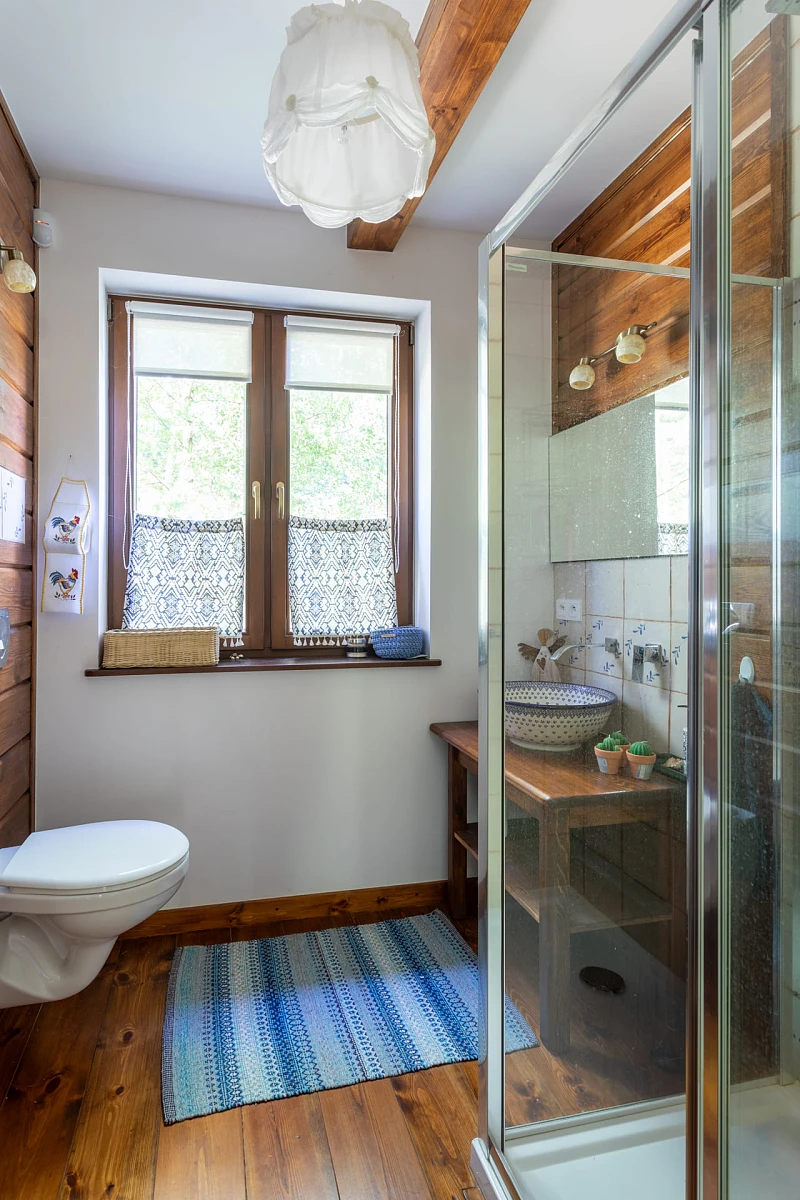 Łazienka w stylu rustykalnym z błękitnymi akcentami w postaci dywanika, przybornika, ale też wzorach umywalki. 