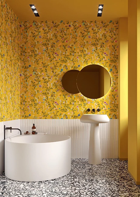 Żółta tapeta z motywem kwiatowym w łazience, kontrastująca z białymi płytkami o nierównej fakturze oraz białą wanną i umywalką wolnostojącą.    